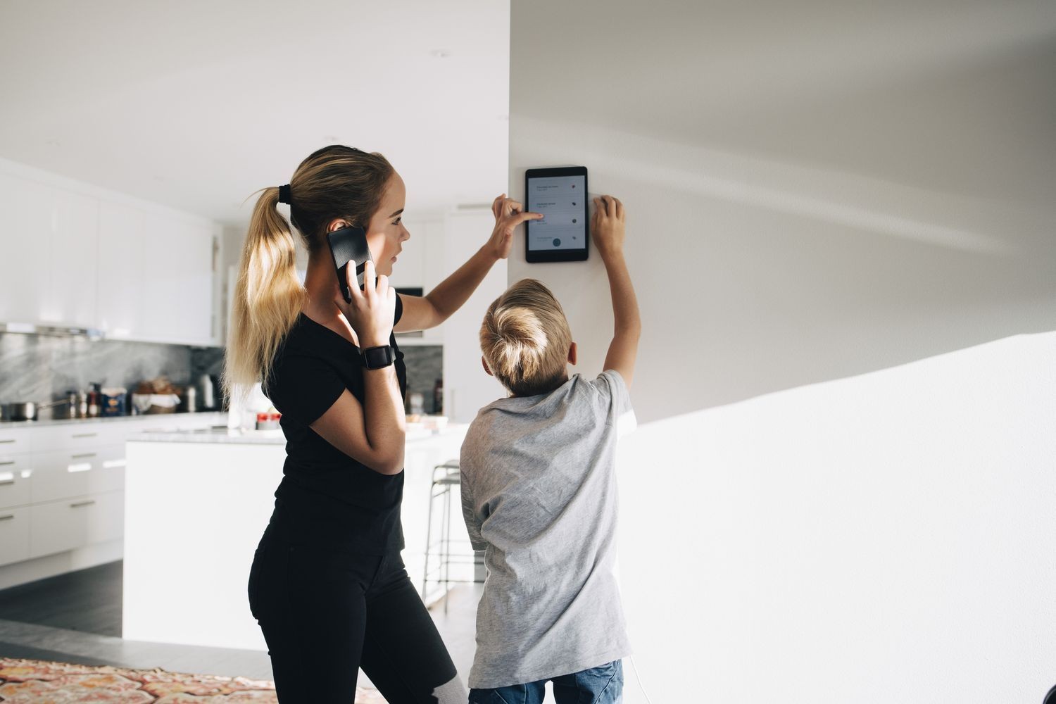 Woman on phone with child setting up a tablet on a wall in a modern kitchen.